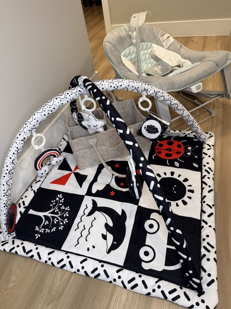 A gray baby bouncer, a colorful baby play mat, and a fully stocked nursery diaper caddy organize station.