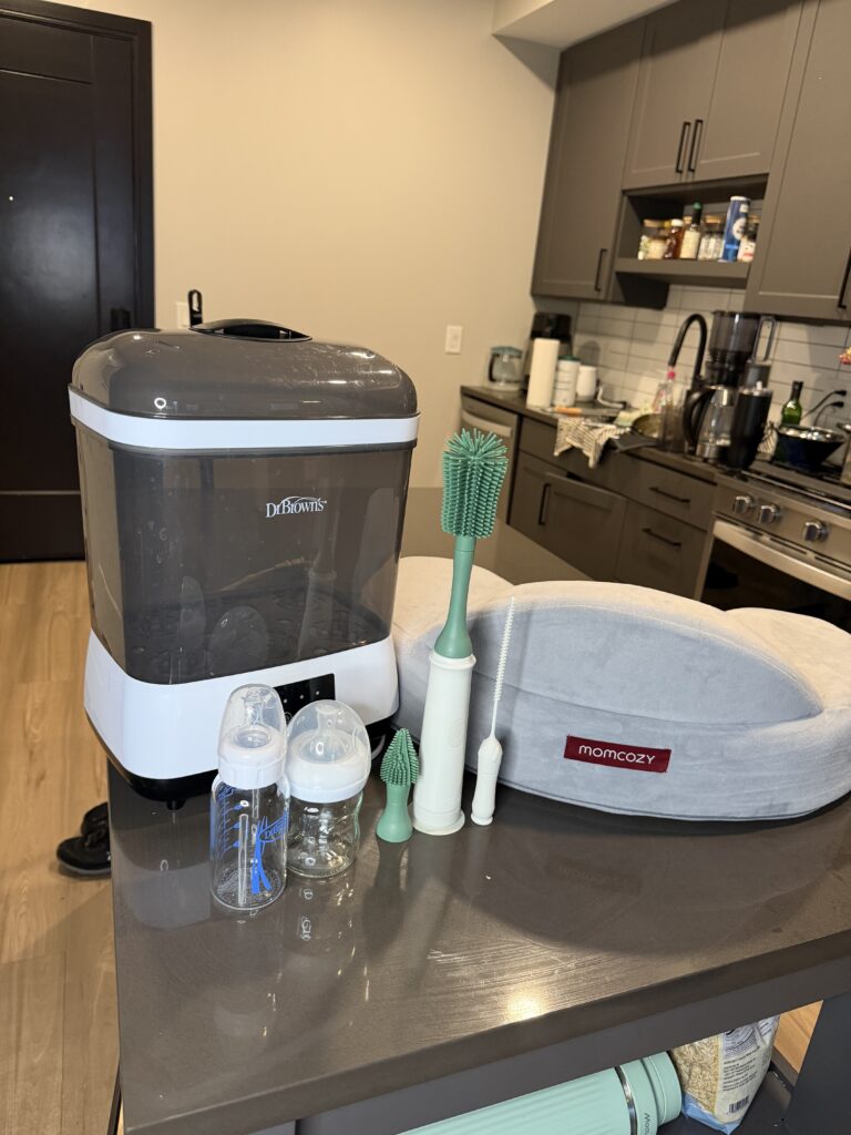 Dr. Brown's bottle sterilizer and electronic brush next to Avent and Dr. Brown's glass baby bottles and a Momcozy firm nursing pillow.