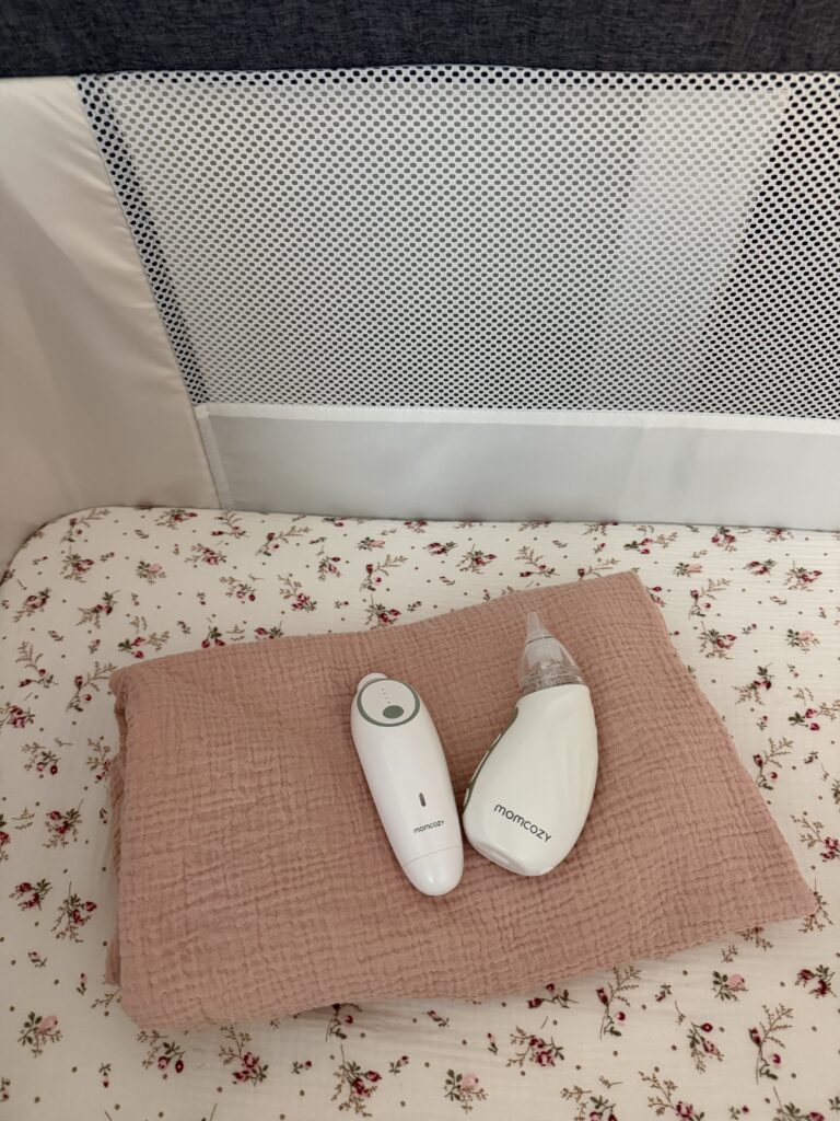 Momcozy nasal aspirator and baby nail trimmer trimmer laying on a white muslin swaddle blanket.